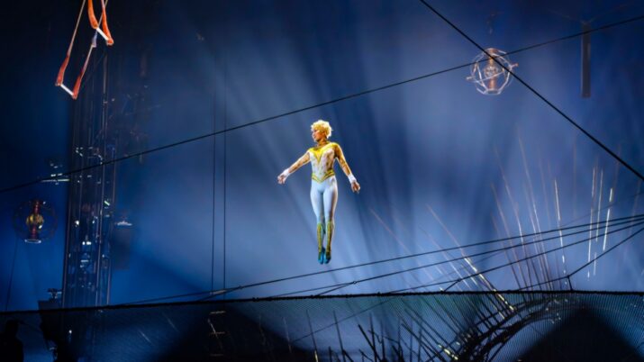 Artista com figurino branco e dourado aparece suspensa no ar sob luzes azuis em um palco de circo.
