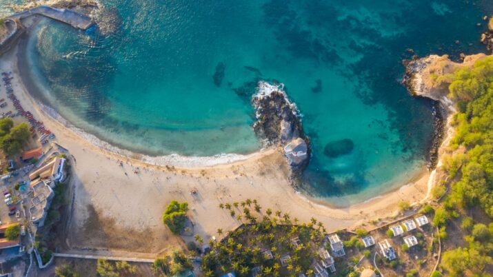 Vista aérea de uma praia em enseada com mar azul-turquesa, faixa de areia e formações rochosas.
