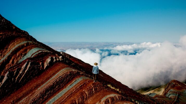 Pessoa de chapéu em pé sobre uma crista de montanha colorida, com nuvens ao fundo e céu azul.