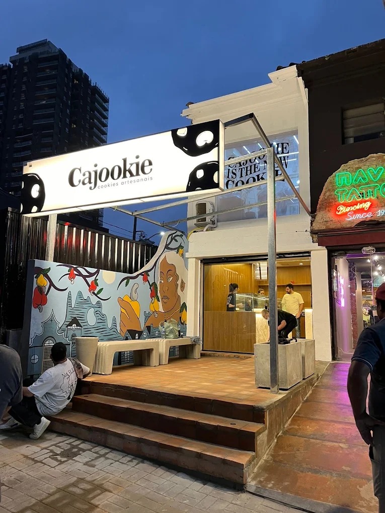 Fachada da loja Cajookie com letreiro iluminado, mural colorido e pessoas na entrada ao anoitecer.