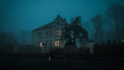 Casa antiga de vários andares cercada por árvores e grade, com janelas iluminadas em meio à neblina ao entardecer.