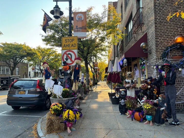 Rua com calçada decorada com bonecos, abóboras e enfeites de Halloween ao lado de carros estacionados.
