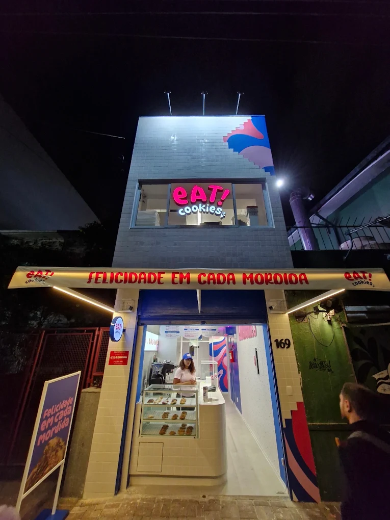 Fachada de uma loja de cookies iluminada à noite, com vitrine, letreiro e uma atendente no balcão.