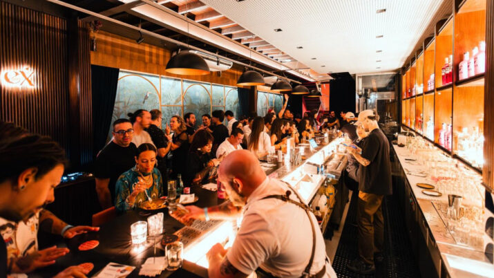Interior de bar lotado, com pessoas sentadas e em pé ao redor de um balcão iluminado.