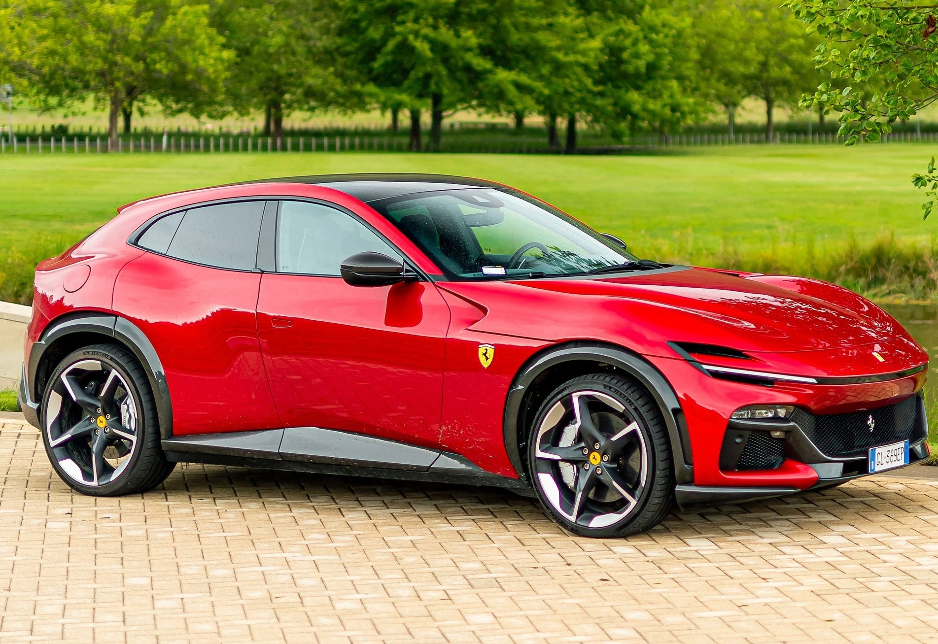 Carro esportivo vermelho da Ferrari estacionado em área externa, com árvores e gramado ao fundo.