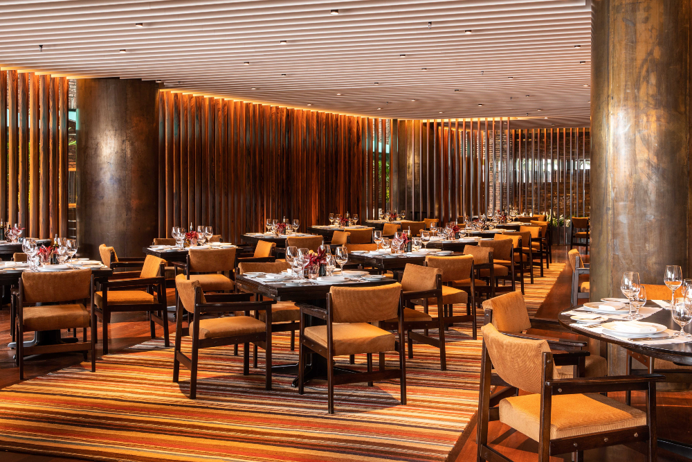 Salão de restaurante com mesas postas, cadeiras estofadas e iluminação quente, sem clientes visíveis.