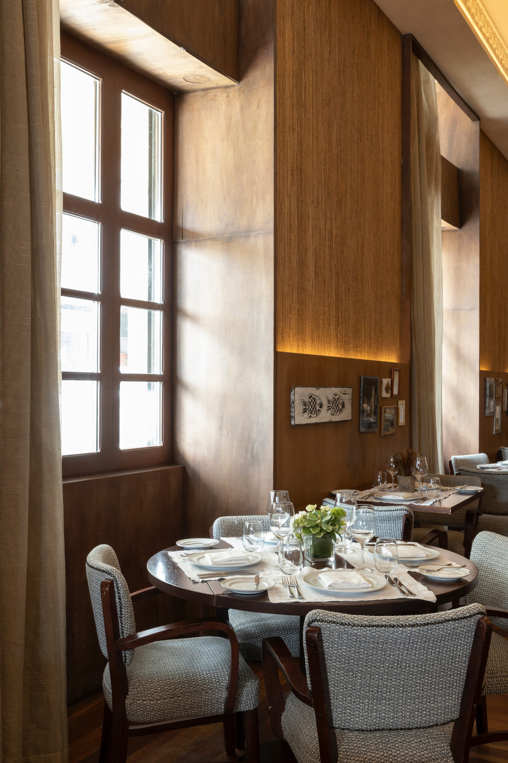 Mesa posta para quatro pessoas em restaurante com paredes de madeira, janelas altas e cadeiras estofadas.