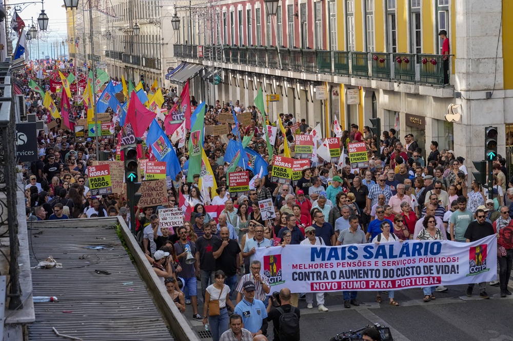 Multidão participa de protesto em rua estreita, com bandeiras coloridas e faixas pedindo mais salário e melhores pensões.
