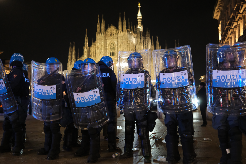 Policiais com capacetes azuis e escudos transparentes com a palavra POLIZIA em uma praça iluminada à noite.