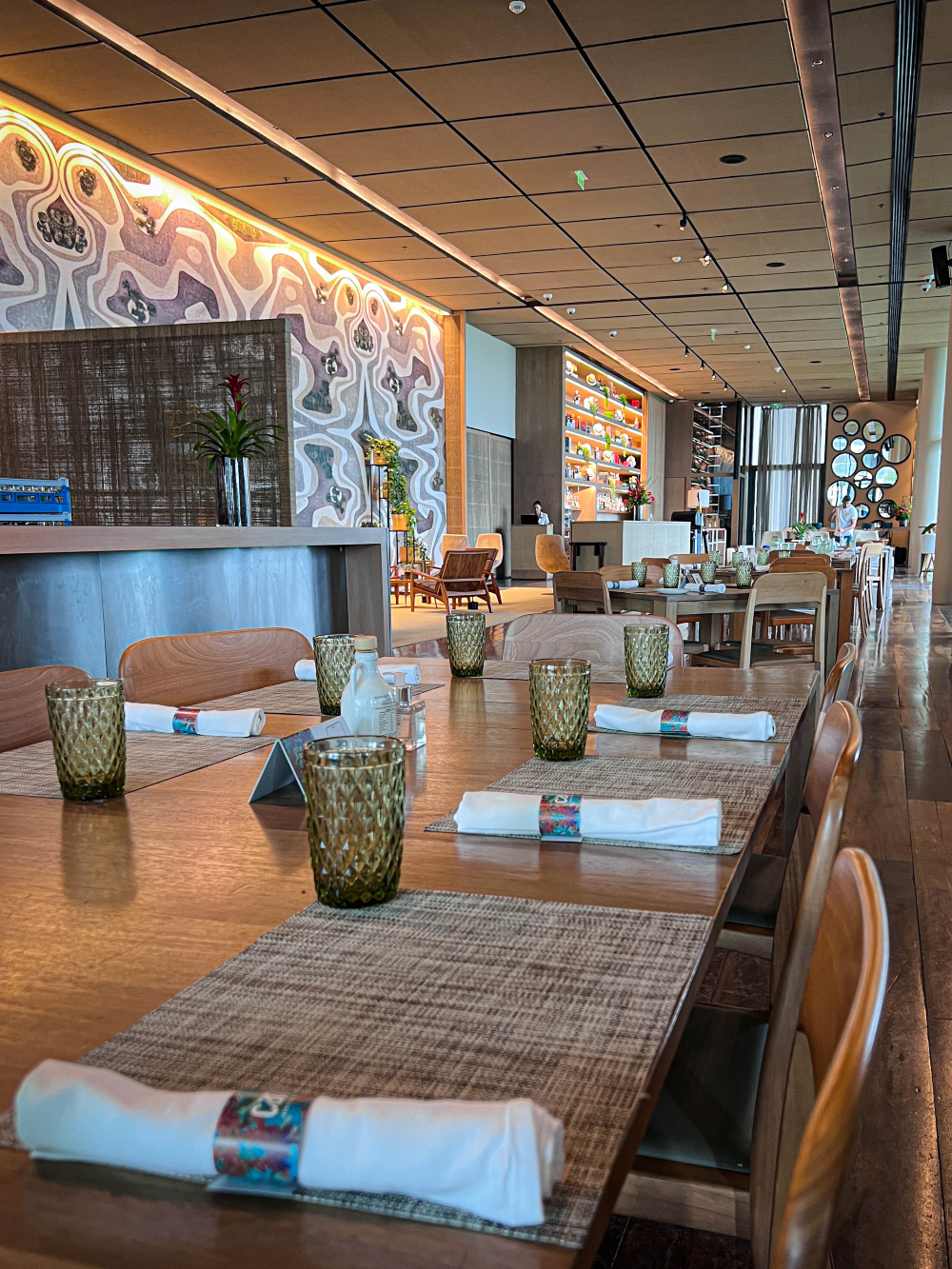 Interior de restaurante com mesas postas, cadeiras de madeira e decoração moderna ao fundo.
