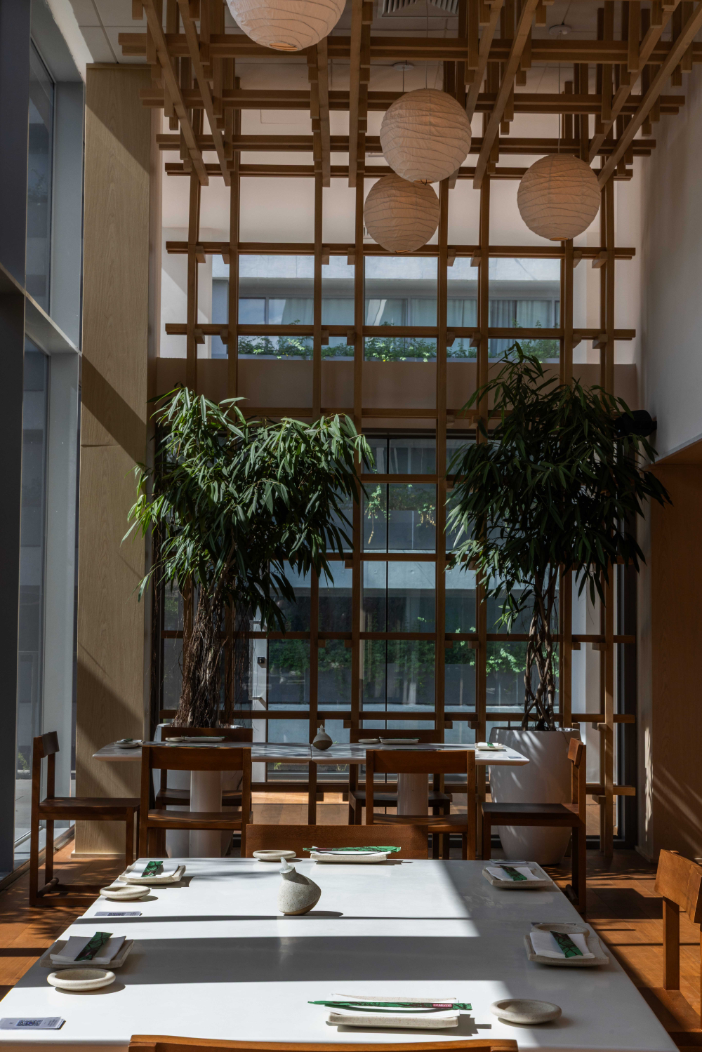 Salão de restaurante com mesas postas, cadeiras de madeira, plantas altas e luminárias pendentes.