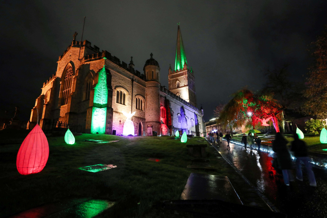 Catedral de pedra iluminada em verde, vermelho e branco à noite, com pessoas caminhando por um caminho molhado ao lado.