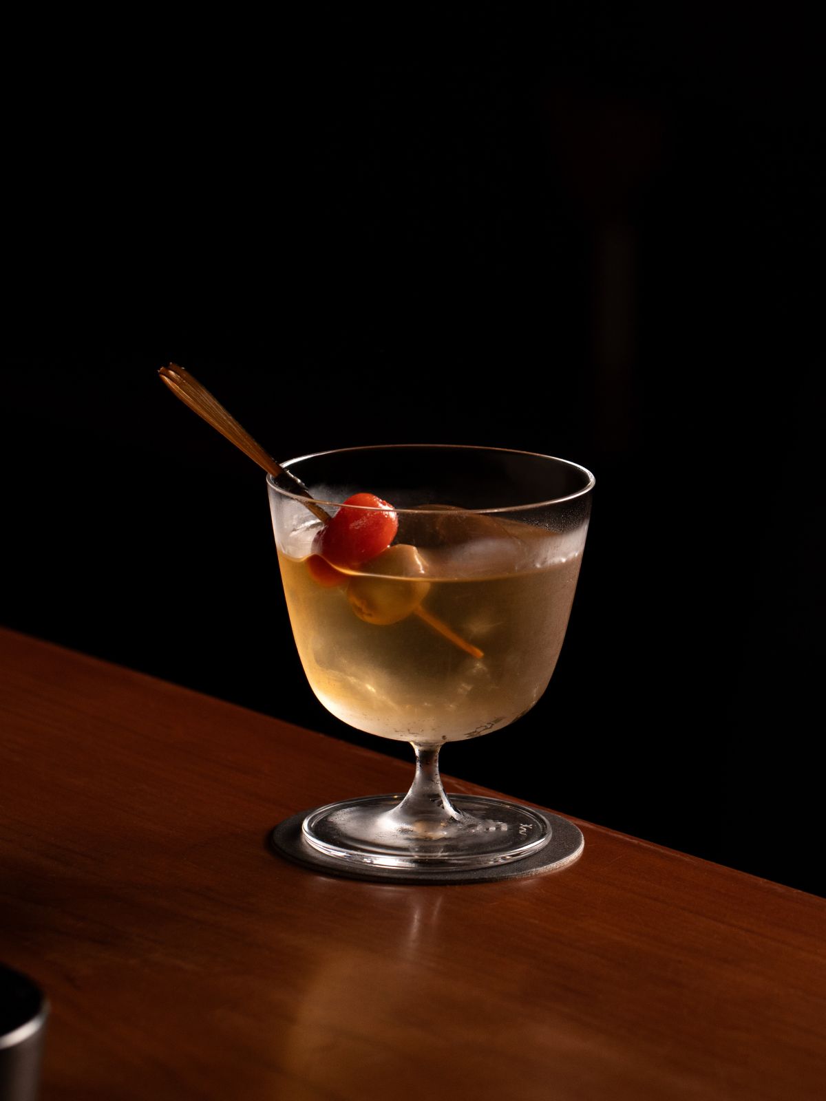 Taça baixa com drink claro, gelo e palito com azeitonas sobre mesa de madeira.