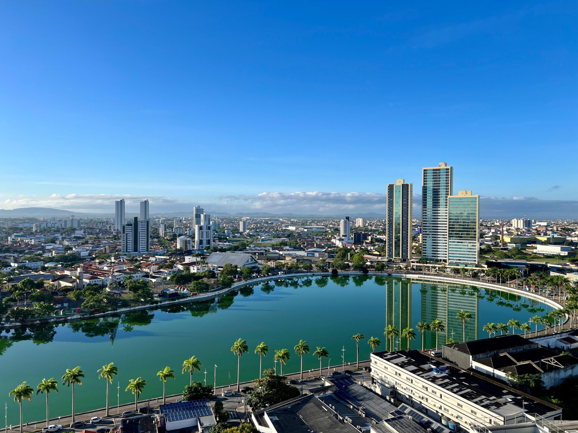 Vista aérea de uma cidade com lago, palmeiras e prédios altos sob céu azul.