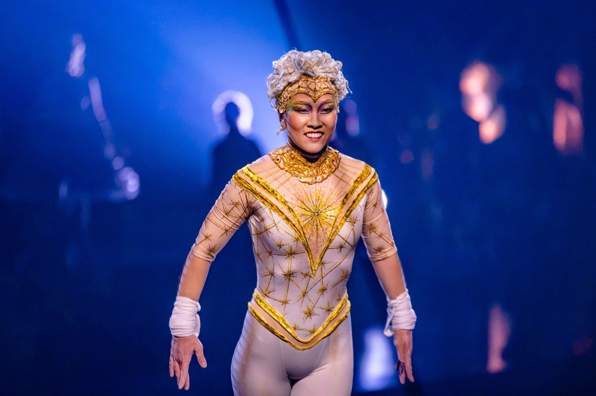 Artista com figurino dourado e maquiagem elaborada sorri em um palco iluminado de azul.
