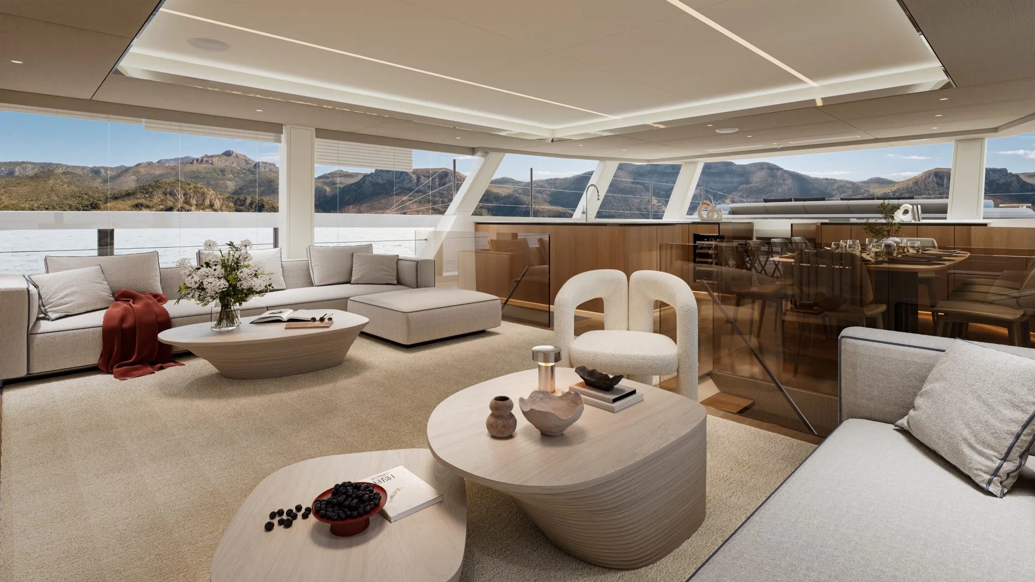 Sala de estar de um iate de luxo com sofás claros, mesas baixas e grandes janelas com vista para o mar e montanhas.