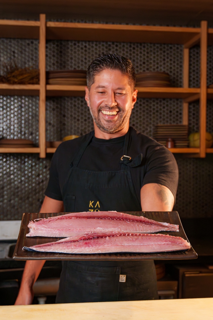 Chef sorridente segura uma bandeja com dois filés de peixe cru em frente a prateleiras de madeira.