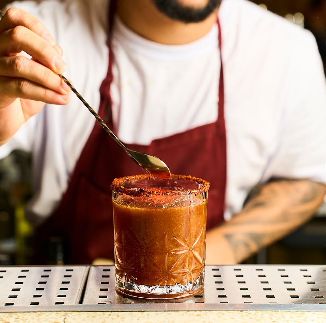 Bartender com avental coloca uma colher sobre um copo com bebida avermelhada e borda temperada.