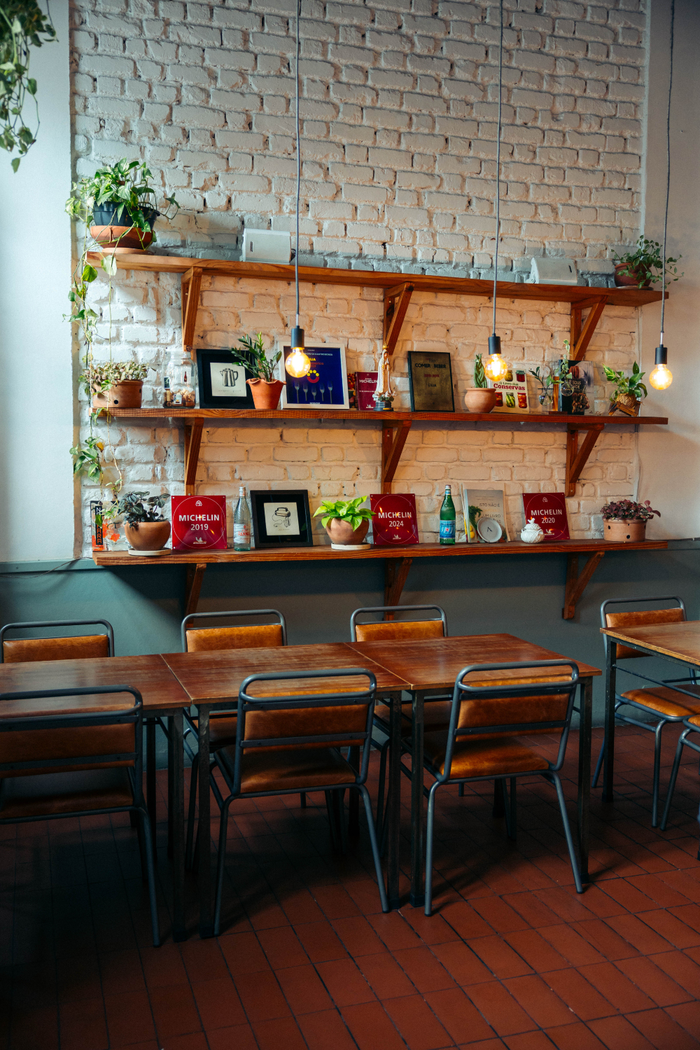 Interior de restaurante com mesas de madeira, cadeiras metálicas, prateleiras com plantas e placas na parede de tijolos.
