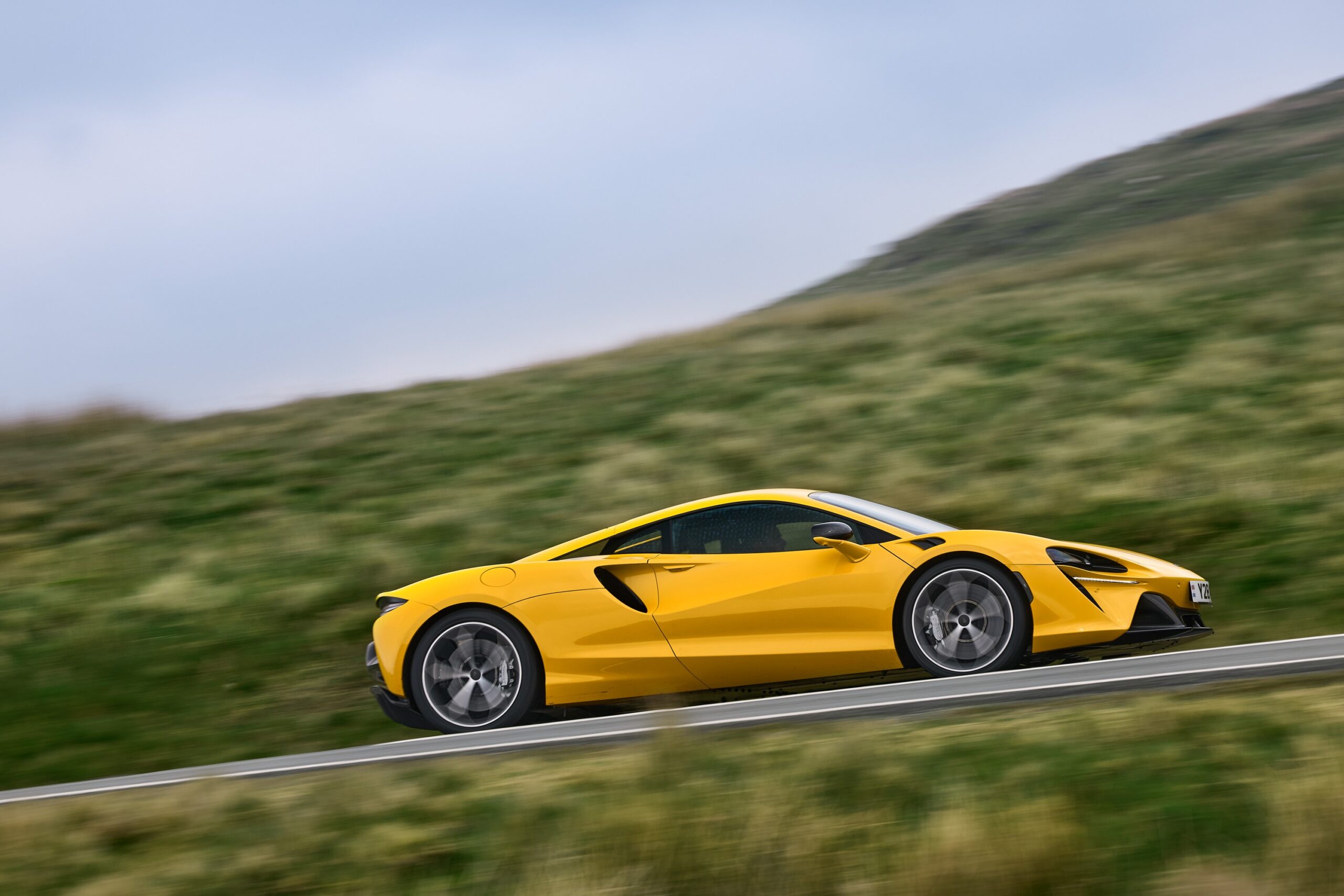 Carro esportivo amarelo trafegando em uma estrada, com fundo desfocado de vegetação e céu aberto.