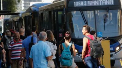 Passageiros aguardam e embarcam em ônibus articulados em um terminal urbano.