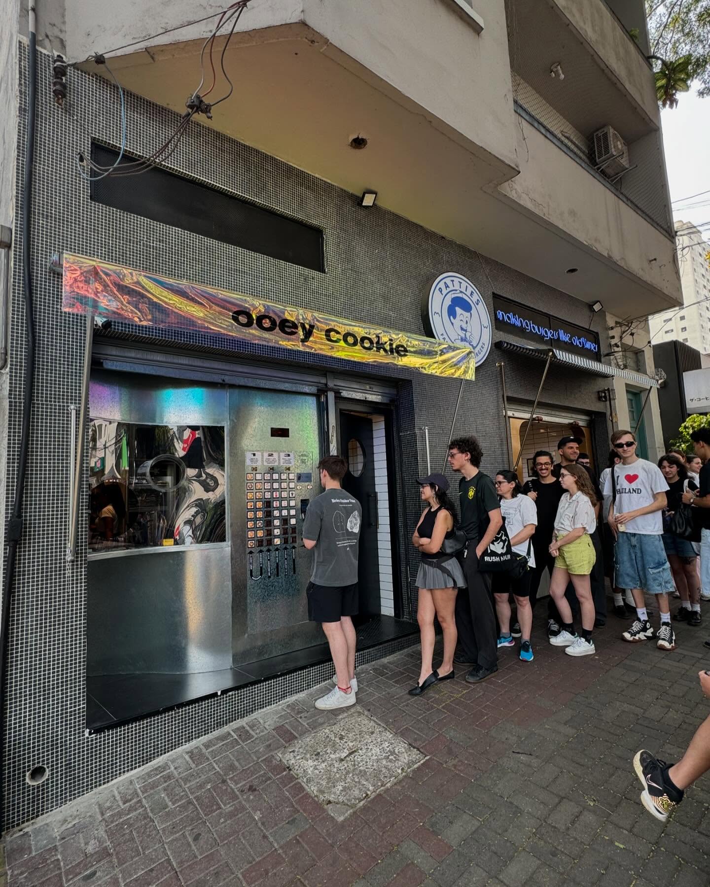 Pessoas aguardam na fila em frente a uma loja de cookies com fachada escura e letreiro colorido.