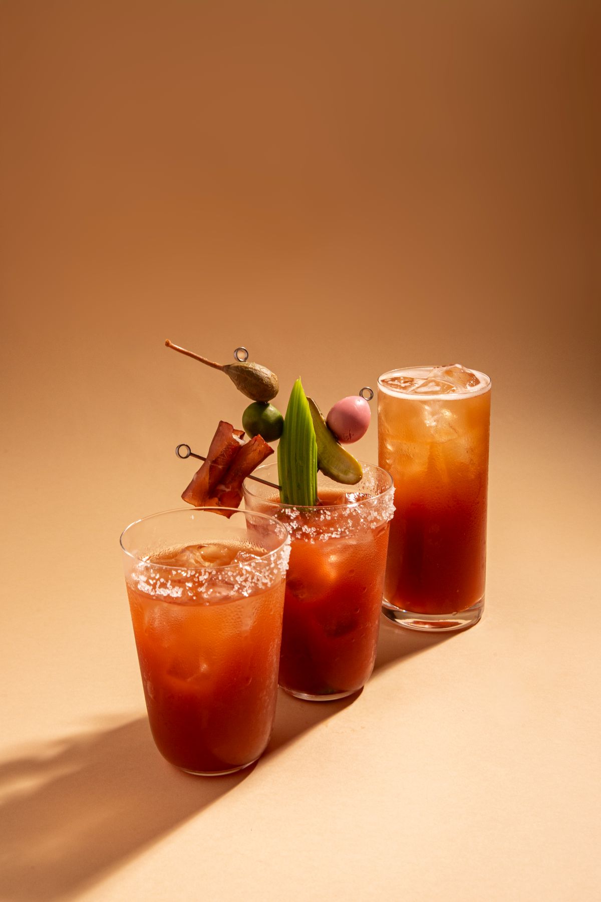 Três copos de drink alaranjado com gelo e borda de sal, decorados com legumes e frutas em fundo bege.