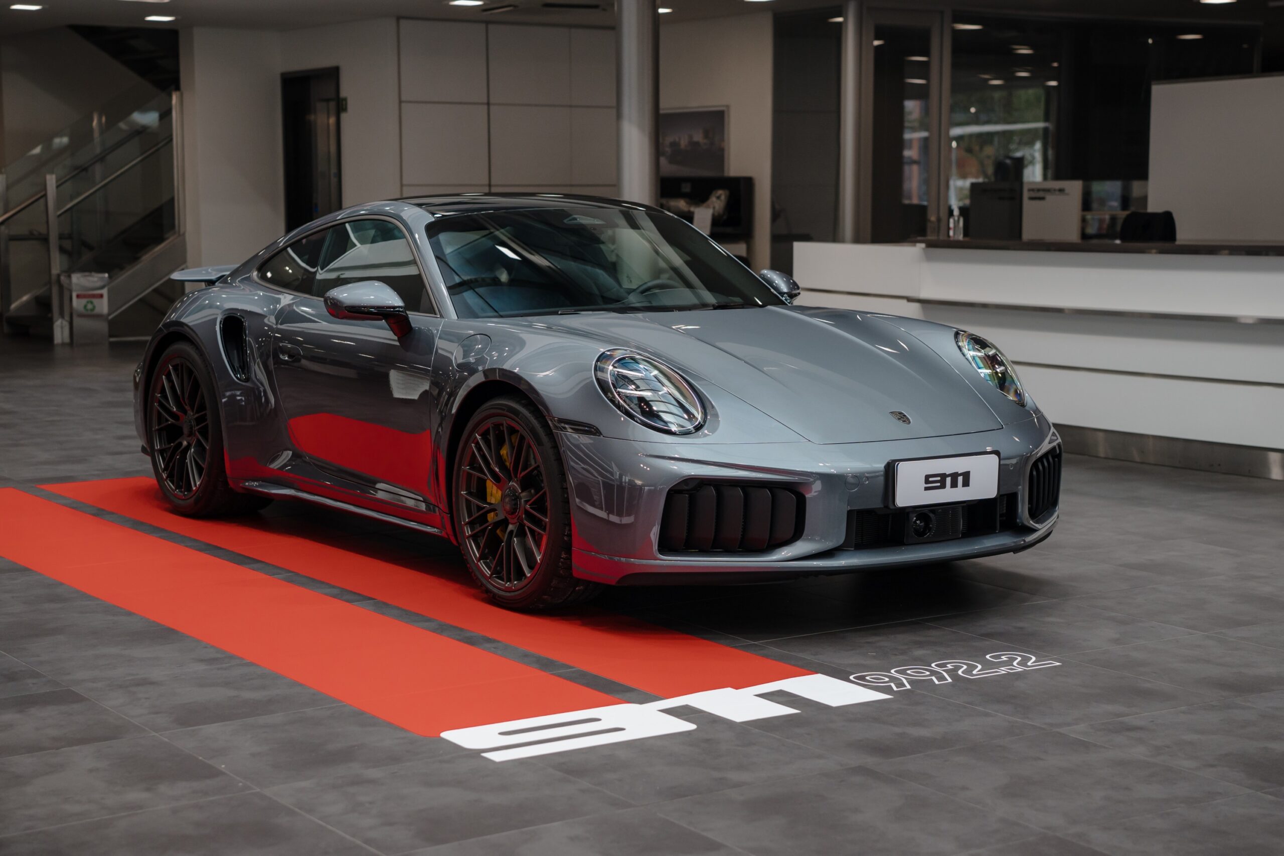 Porsche 911 cinza exibido em uma concessionária, sobre faixa vermelha no piso.