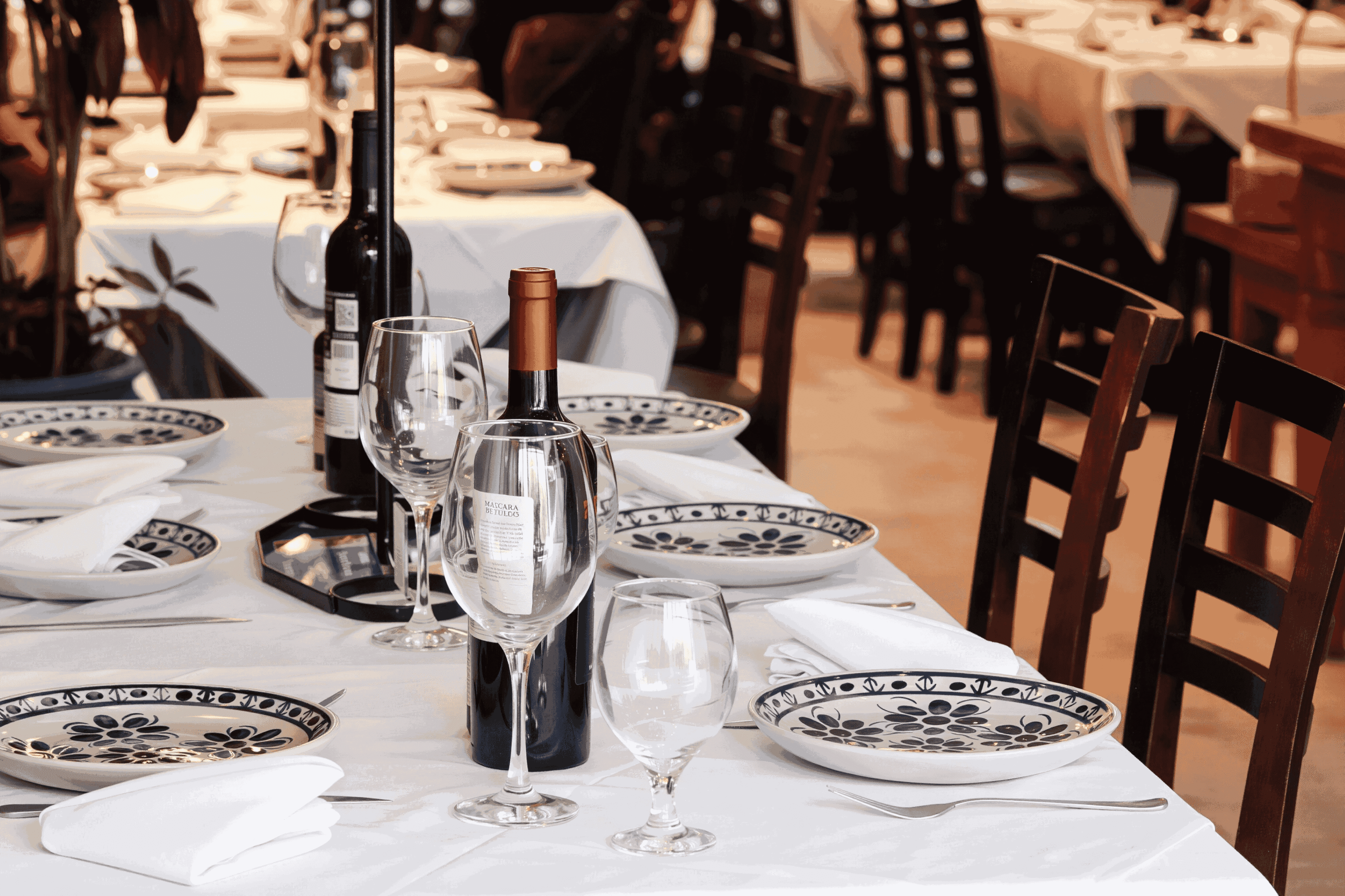 Mesa posta em restaurante com taças, pratos, guardanapos e garrafas de vinho.
