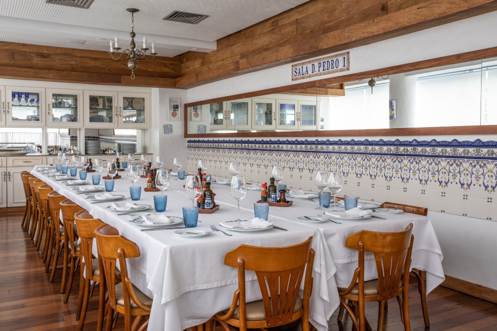 Salão de restaurante com mesa longa posta para várias pessoas, cadeiras de madeira e decoração em azulejos azuis.