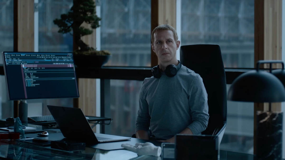 Homem sentado em uma cadeira diante de uma mesa com notebook e monitor, em um escritório moderno com vista para janelas grand