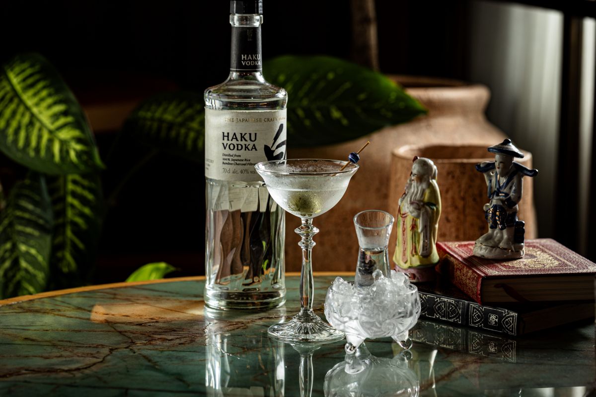 Garrafa de vodka, taça de coquetel e copo com gelo sobre uma mesa, ao lado de pequenas estátuas decorativas.