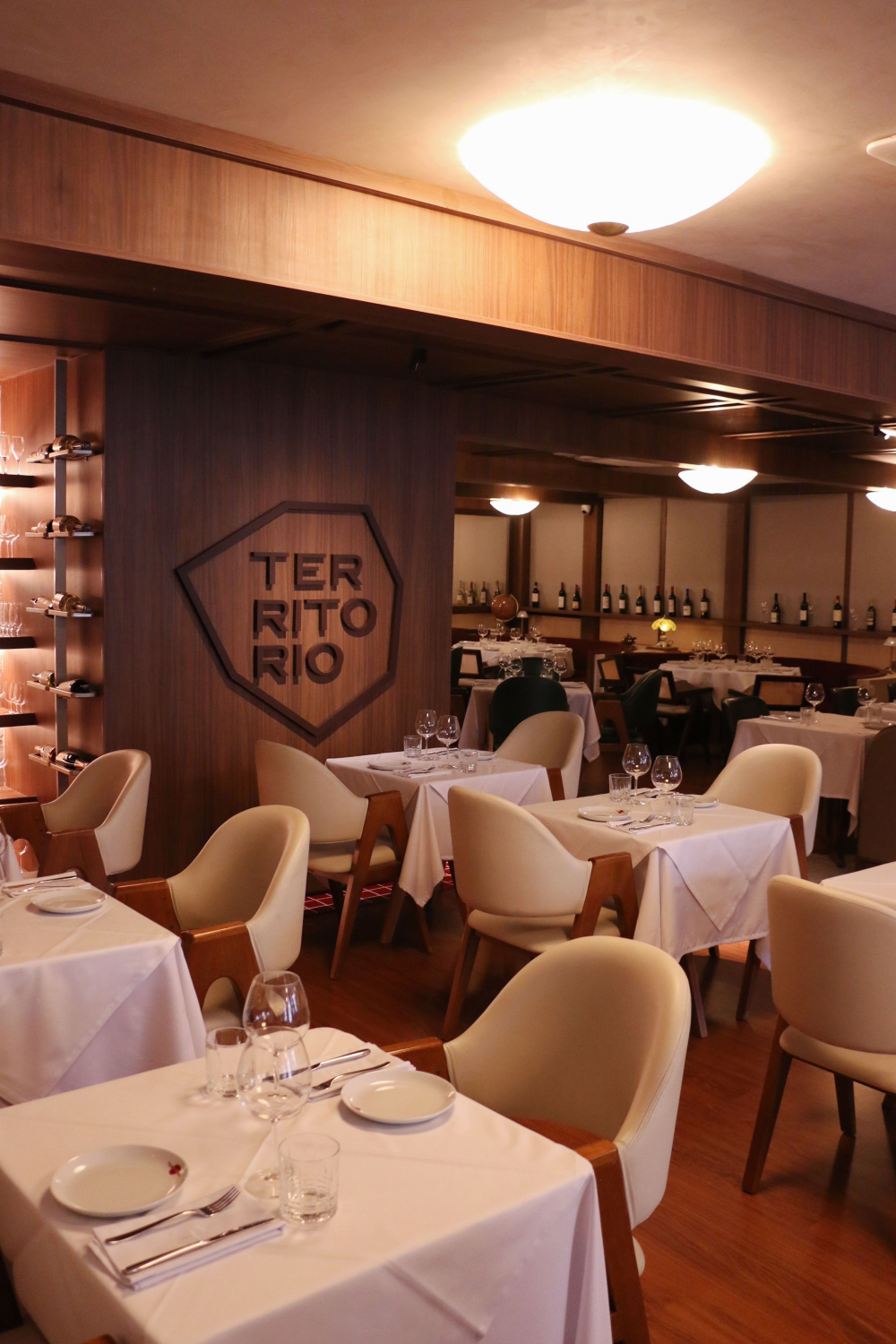 Interior de restaurante com mesas postas, cadeiras claras e iluminação amarelada, com o nome Território em uma parede de made
