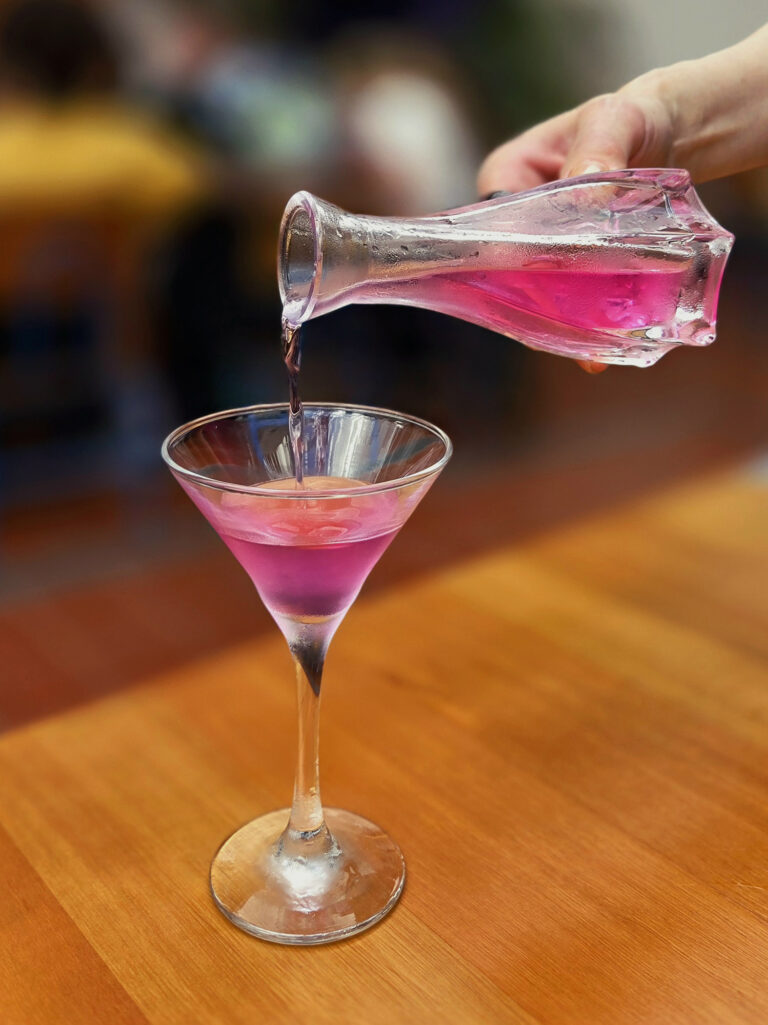 Mão despeja bebida rosa de uma garrafa em uma taça de coquetel sobre mesa de madeira.