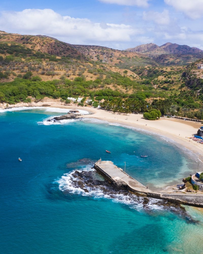 Vista aérea de uma praia em enseada, com mar azul, faixa de areia, píer de pedra e morros ao fundo.