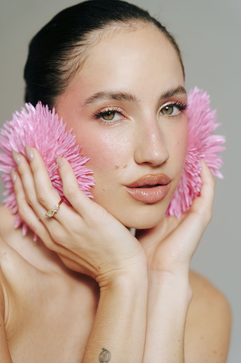 Mulher em close com flores rosas nas mãos ao lado do rosto e cabelo preso para trás.