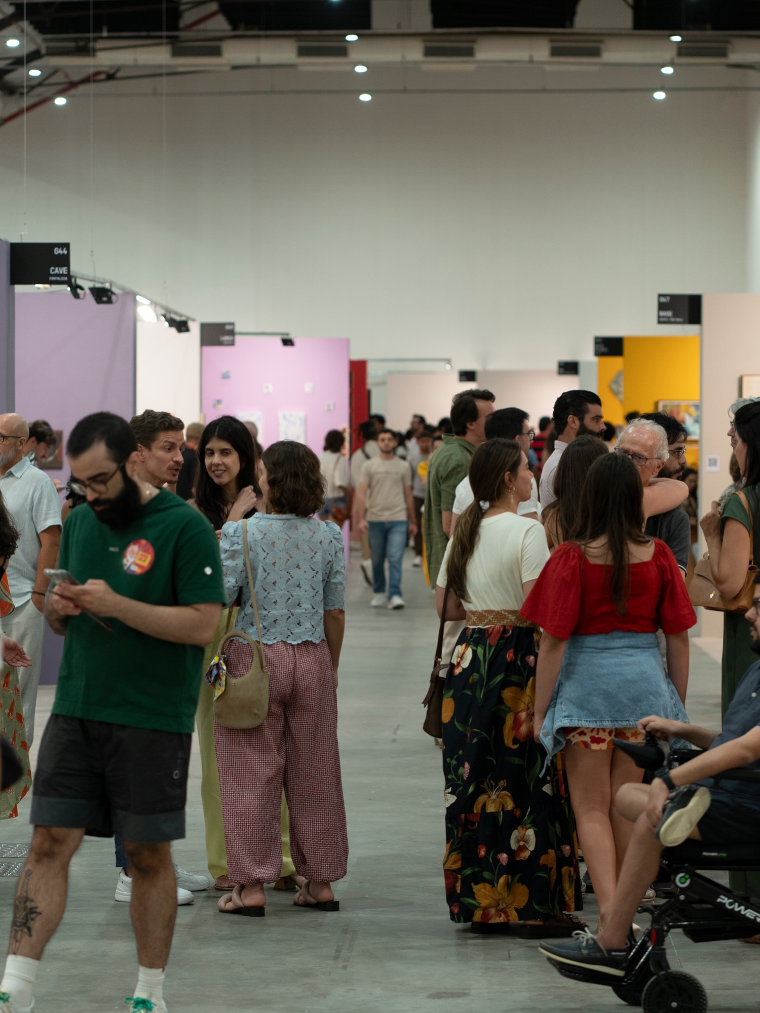 Pessoas circulam por um pavilhão de feira, entre estandes coloridos e obras expostas ao fundo.