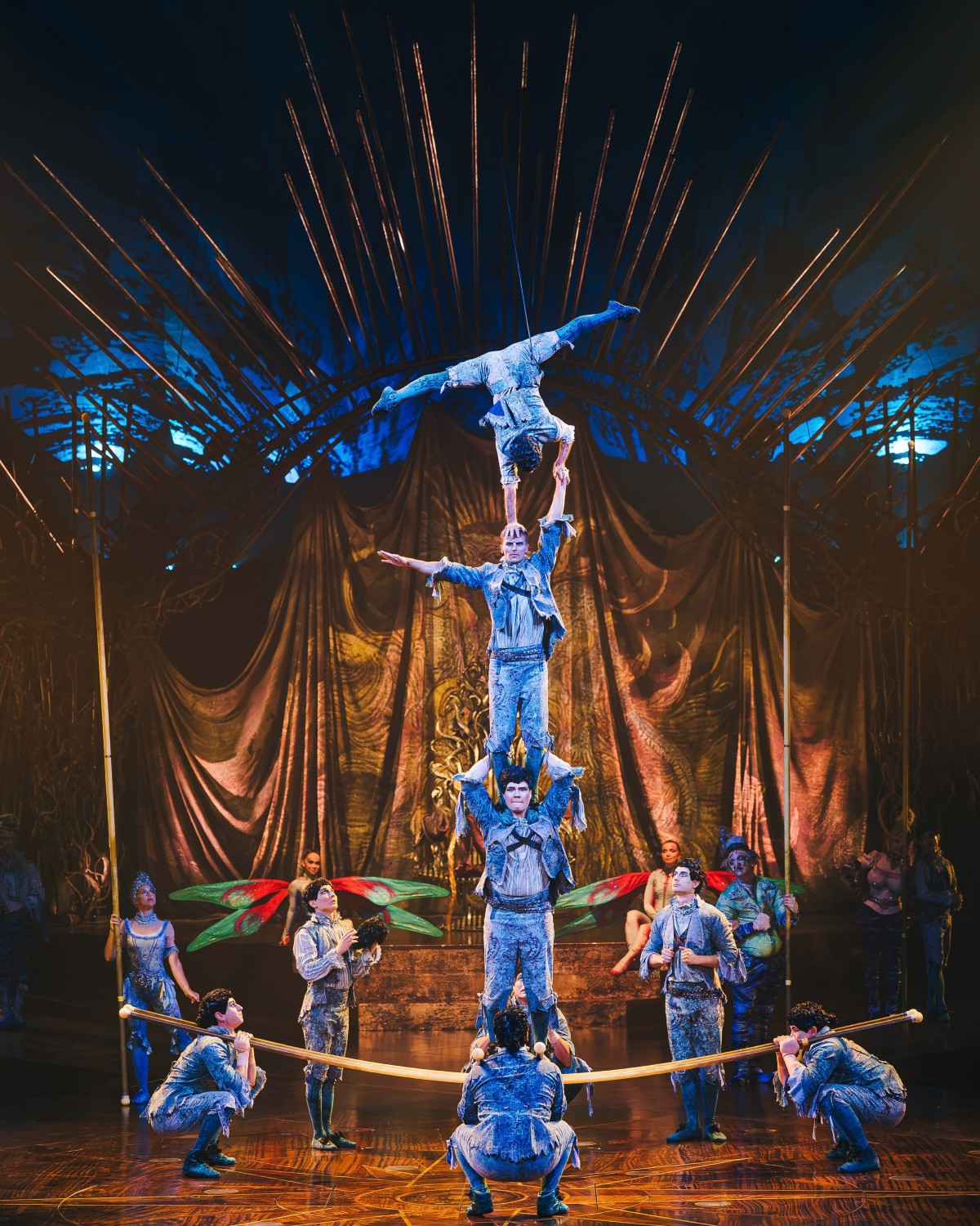 Grupo de acrobatas em figurinos azuis faz uma pirâmide humana em um palco iluminado.