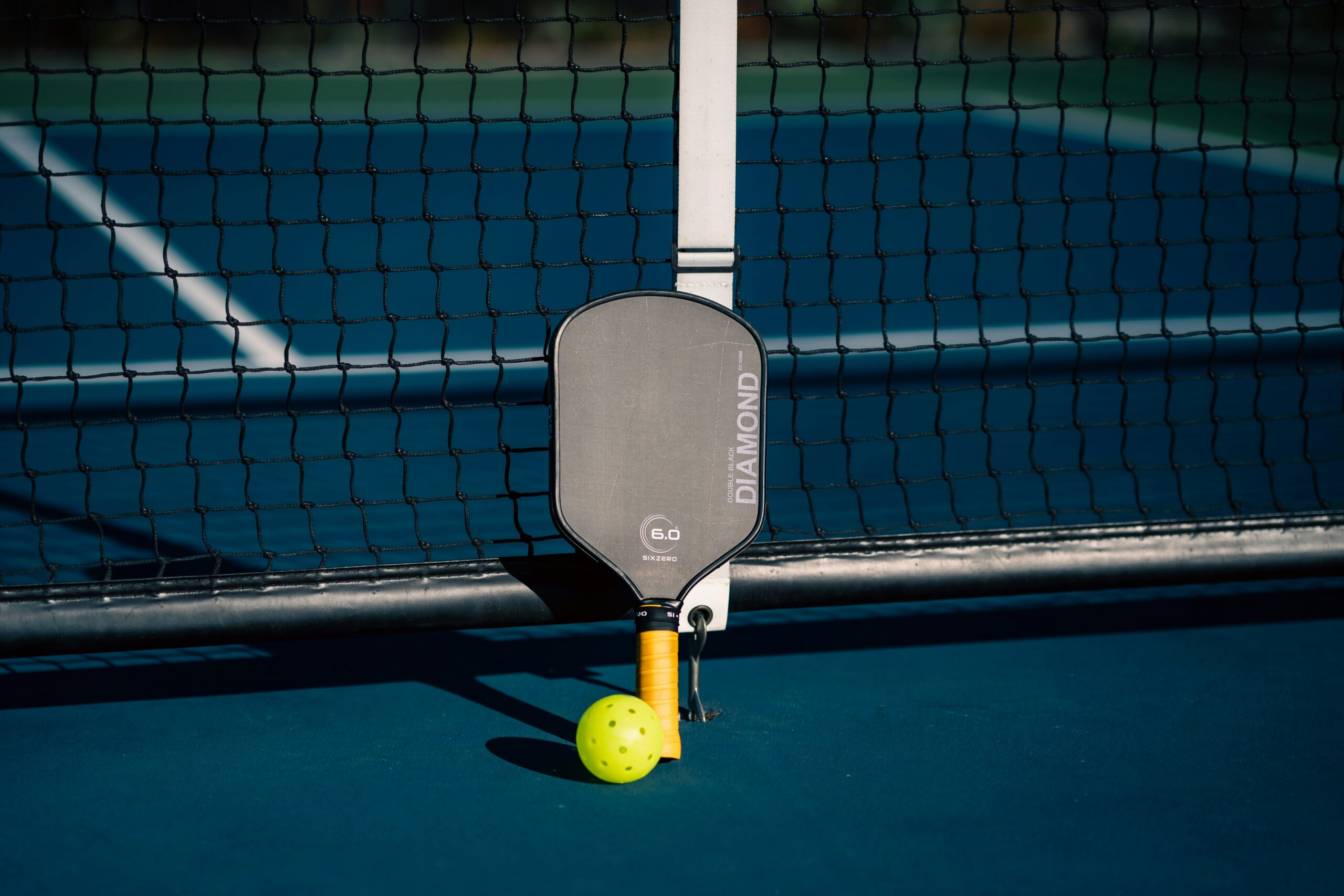 Raquete de pickleball encostada na rede, com uma bola amarela no chão da quadra azul.