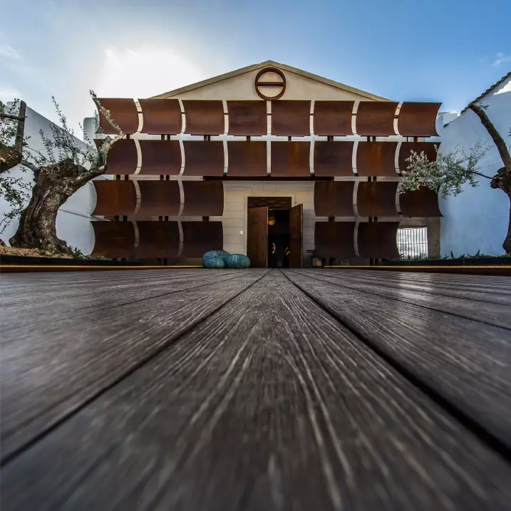 Fachada de um prédio com painéis marrons curvos e porta central, vista de baixo sobre um deck de madeira.