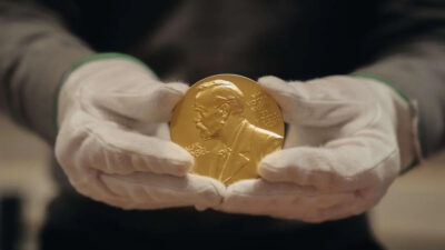 Mãos com luvas brancas seguram uma medalha dourada do Nobel.