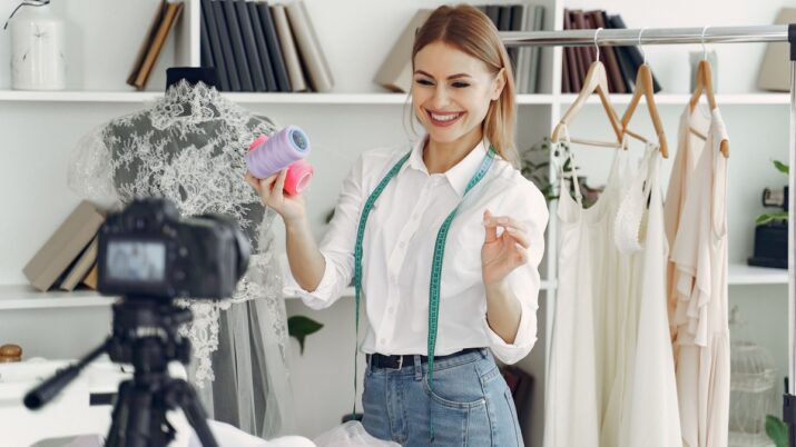 Mulher sorrindo em estúdio de moda, com fita métrica no pescoço, segurando carretéis de linha ao lado de um manequim e roupas