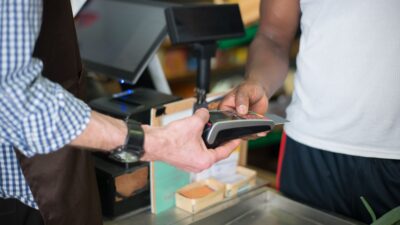 Pessoa segura uma maquininha de cartão enquanto outra aproxima o cartão para pagamento no balcão.