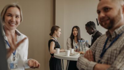 Pessoas conversam em pé ao redor de uma mesa alta com copos, garrafas de água e petiscos.