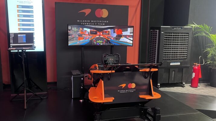 Simulador de corrida com cockpit da McLaren, volante e monitor exibindo uma pista, em ambiente de exposição da Mastercard.