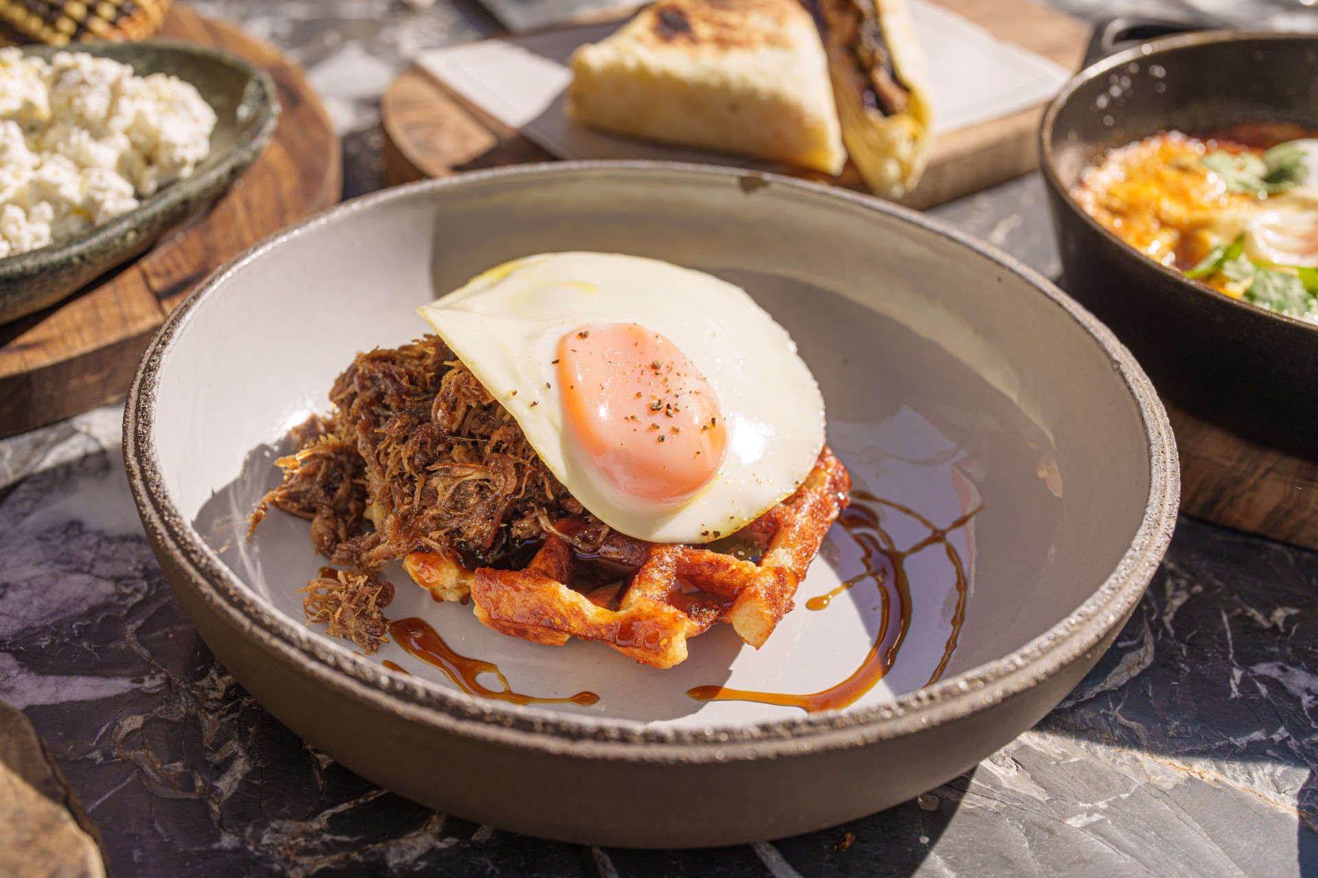 Prato fundo com waffle, carne desfiada e ovo frito, com outros pratos ao fundo.