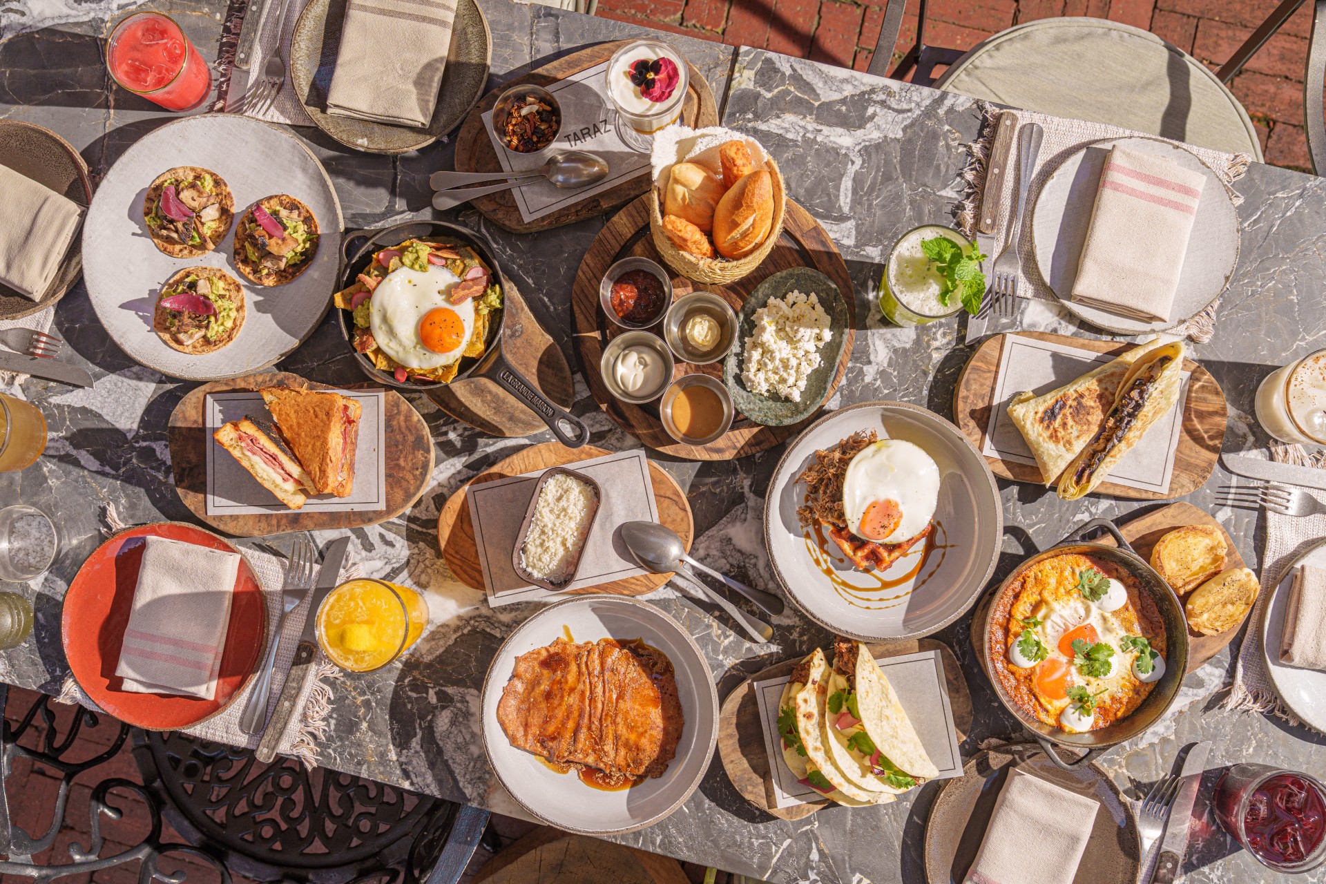 Mesa vista de cima com pratos de café da manhã, pães, ovos, bebidas e talheres dispostos em várias posições.