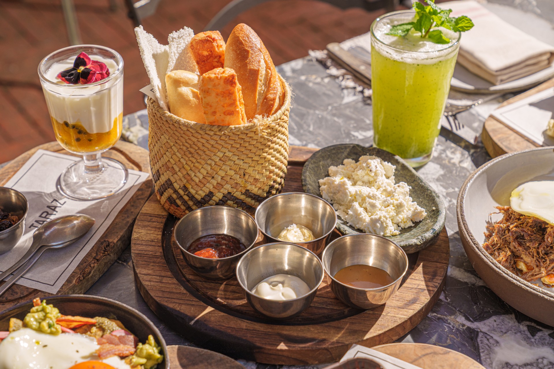 Mesa com cesta de pães, copos de suco e iogurte, potes com acompanhamentos e pratos de café da manhã.